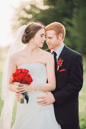 A romantic wedding couple embraces outdoors at sunset with the bride holding red roses and wearing a white veil.