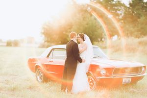 A vintage red Ford Mustang parked in a sunlit field with a couple in formal attire embracing beside it.