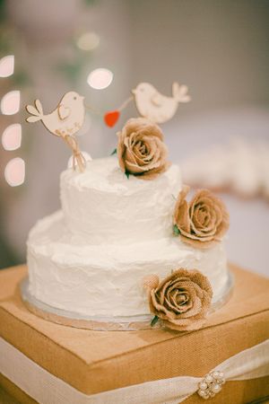 White two-tier wedding cake decorated with gold roses and cute bird cake toppers against a soft bokeh background.