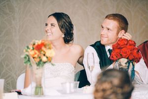A couple sits at their wedding reception table with orange and red floral centerpieces on display.