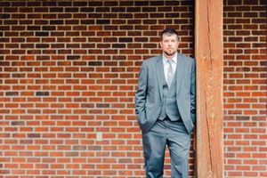 A person in a gray suit stands against a rustic brick wall with wooden beam, giving a formal business portrait.
