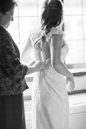 A bride in a white lace wedding dress is being helped with final adjustments before the ceremony in a sunlit room.