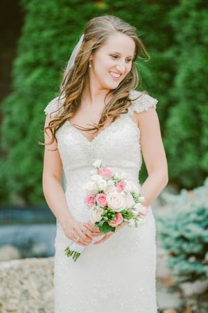 A bride in a lace wedding dress poses in front of lush greenery while holding a pink and white rose bouquet.