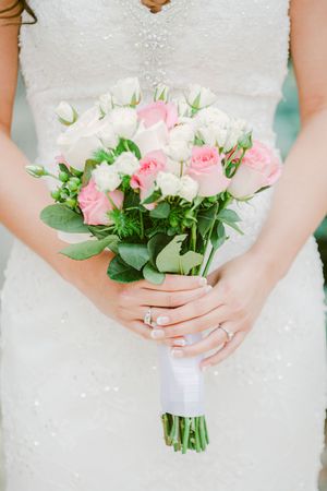 A bridal bouquet of pink and white roses with greenery wrapped in white ribbon, held against a sparkly wedding dress.