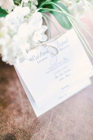 Elegant white wedding invitation card with blue text hangs beside white flowers on a rustic textured surface.