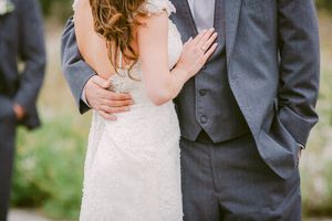 Close-up photo showing formal wedding attire with a white lace dress and charcoal gray suit during outdoor ceremony.
