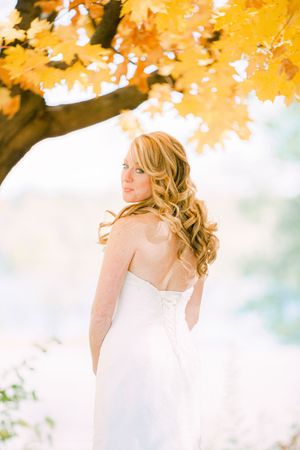 A bride in a white strapless gown stands beneath golden autumn maple leaves on a bright fall day.