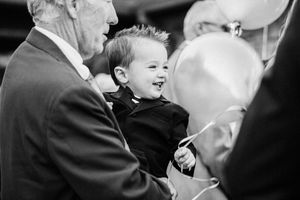A joyful young child laughs while holding a balloon at a gathering with adults in black and white photography.
