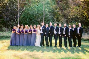 Wedding party in purple dresses and black tuxedos lined up outdoors against autumn foliage background.