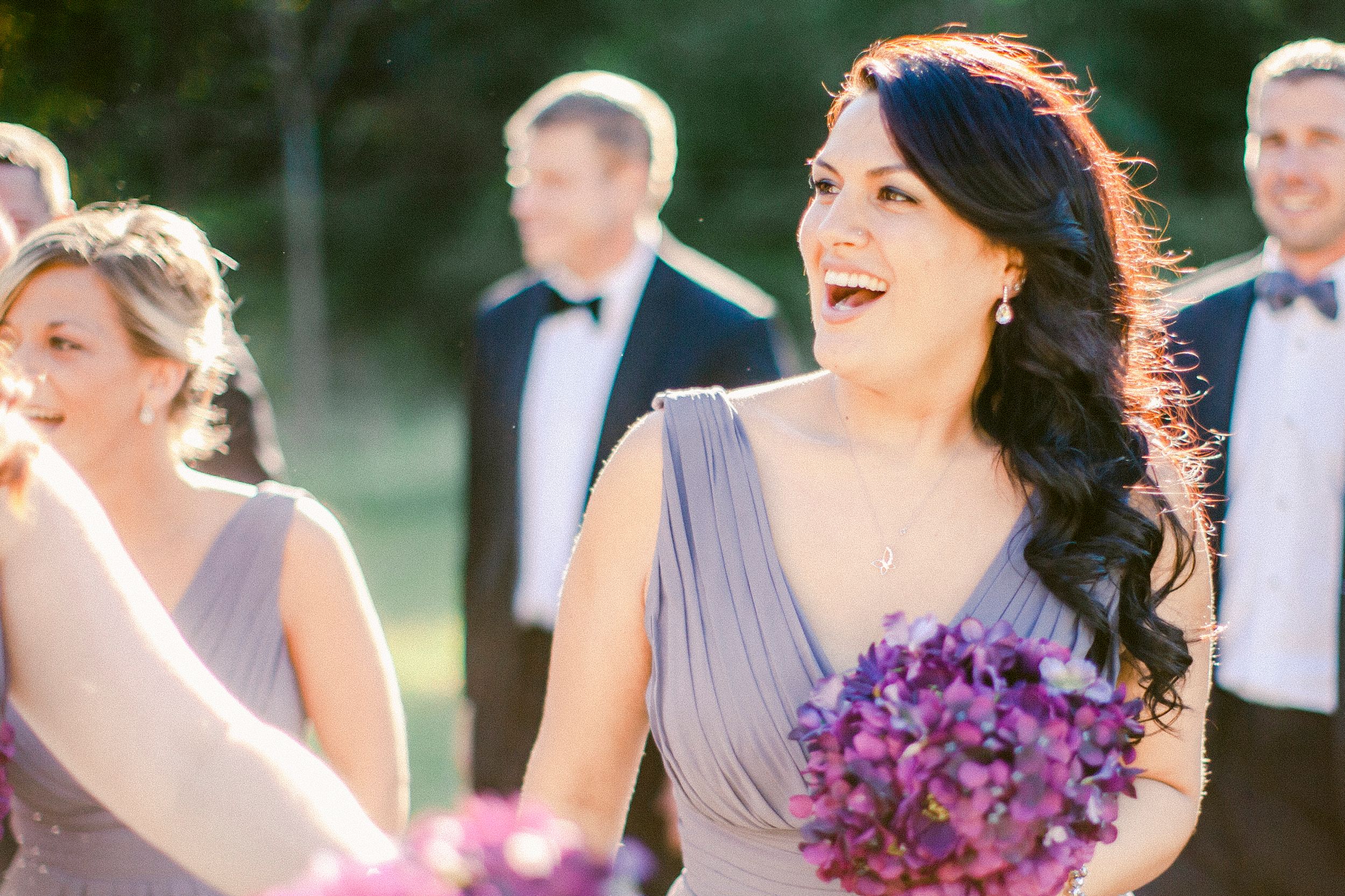 Wedding party members in formal attire share a joyful moment with vibrant purple bouquets during outdoor celebration.