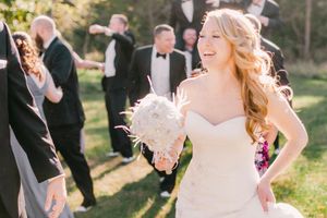 A bride in a white dress holding a white bouquet poses in sunlit outdoor wedding ceremony setting with guests in background.