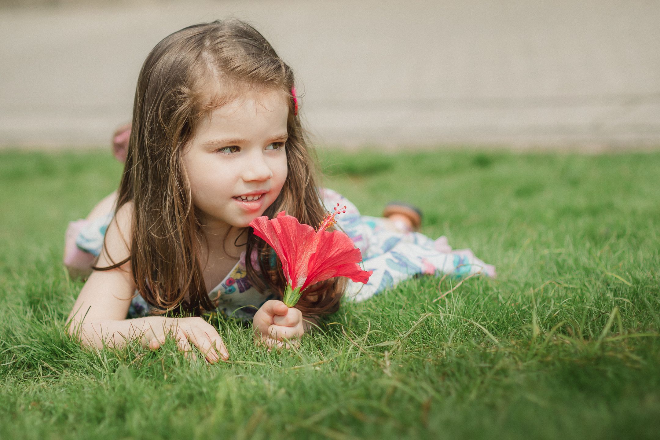 Young child lying on green grass holding a bright red flower in a spring or summer setting.