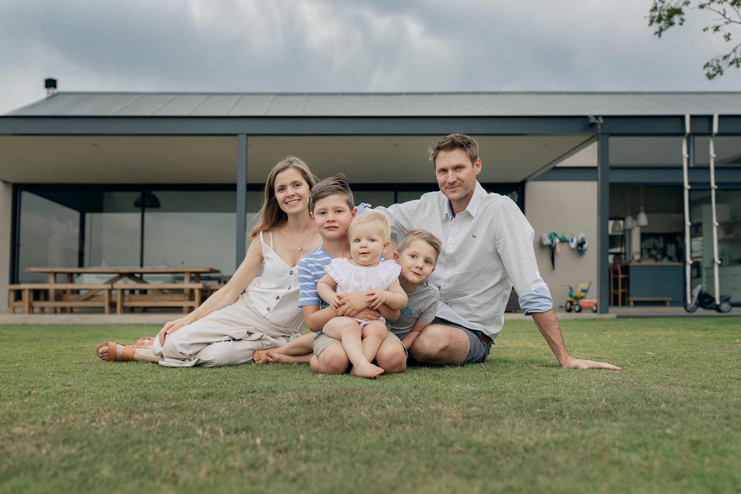 Family relaxing together on lawn in front of modern house with covered patio.