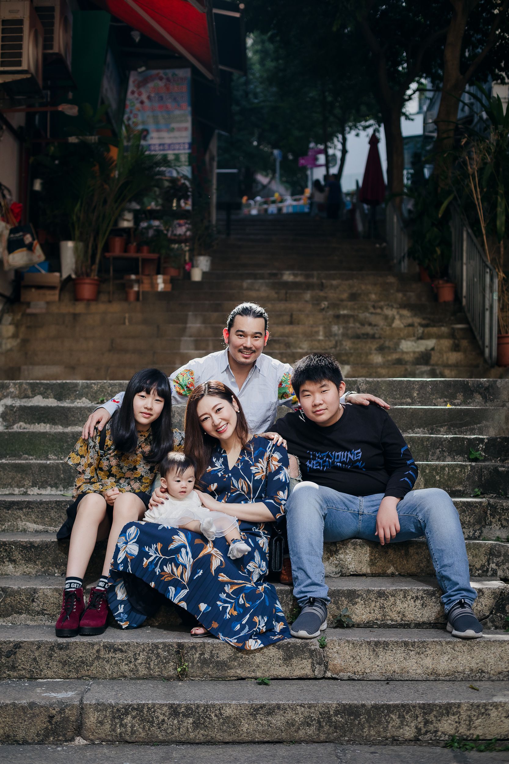 A diverse group sits together on stone steps surrounded by plants and urban scenery.