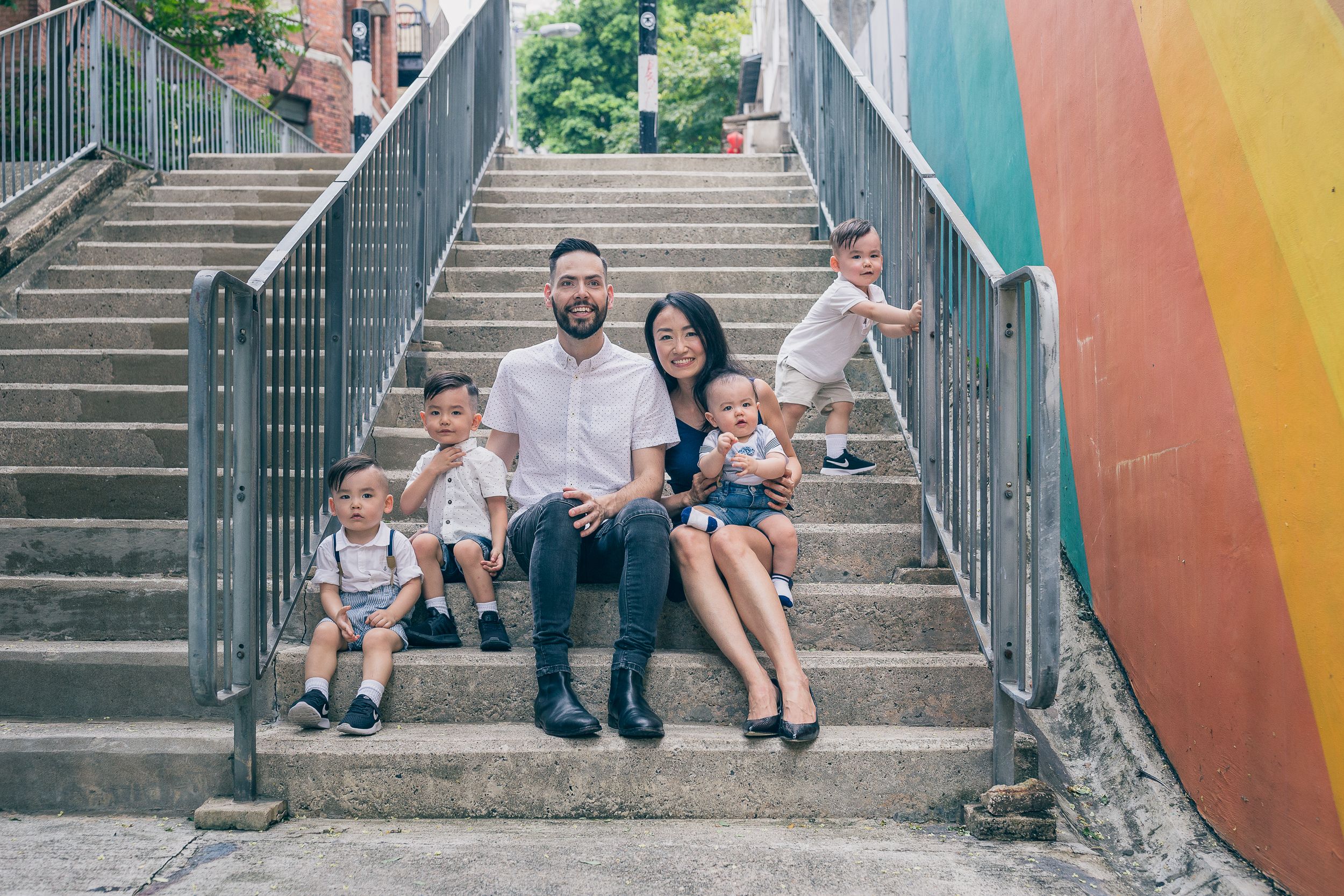 A family sits on colorful urban stairs with a rainbow painted wall in the background.
