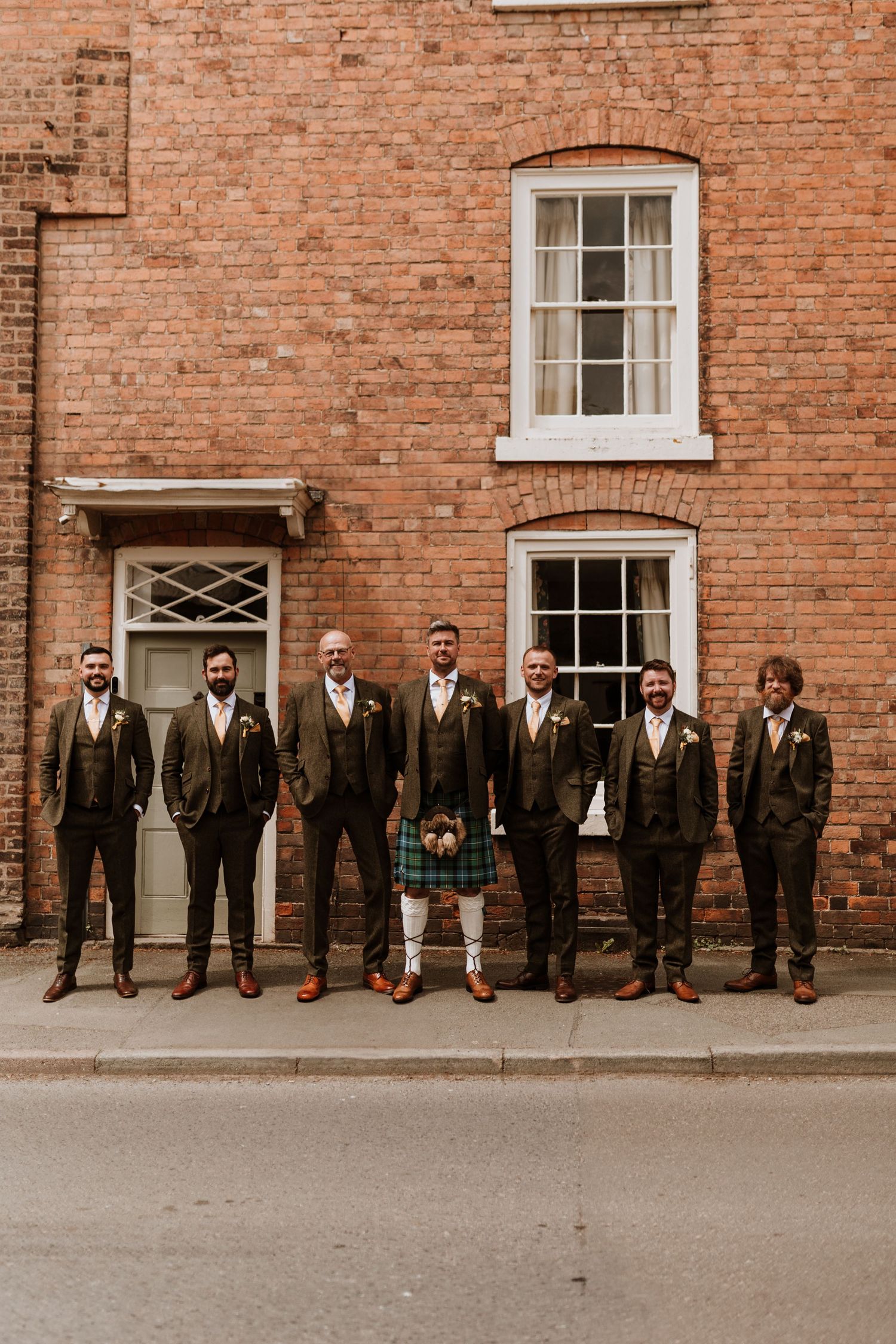 Wedding party in vintage brown suits stands in line against rustic brick wall.