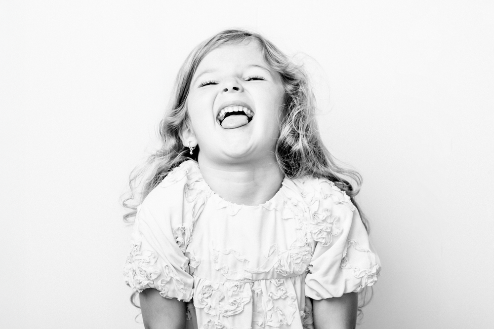 A child with curly hair wearing a white lace dress laughs joyfully against a white background.