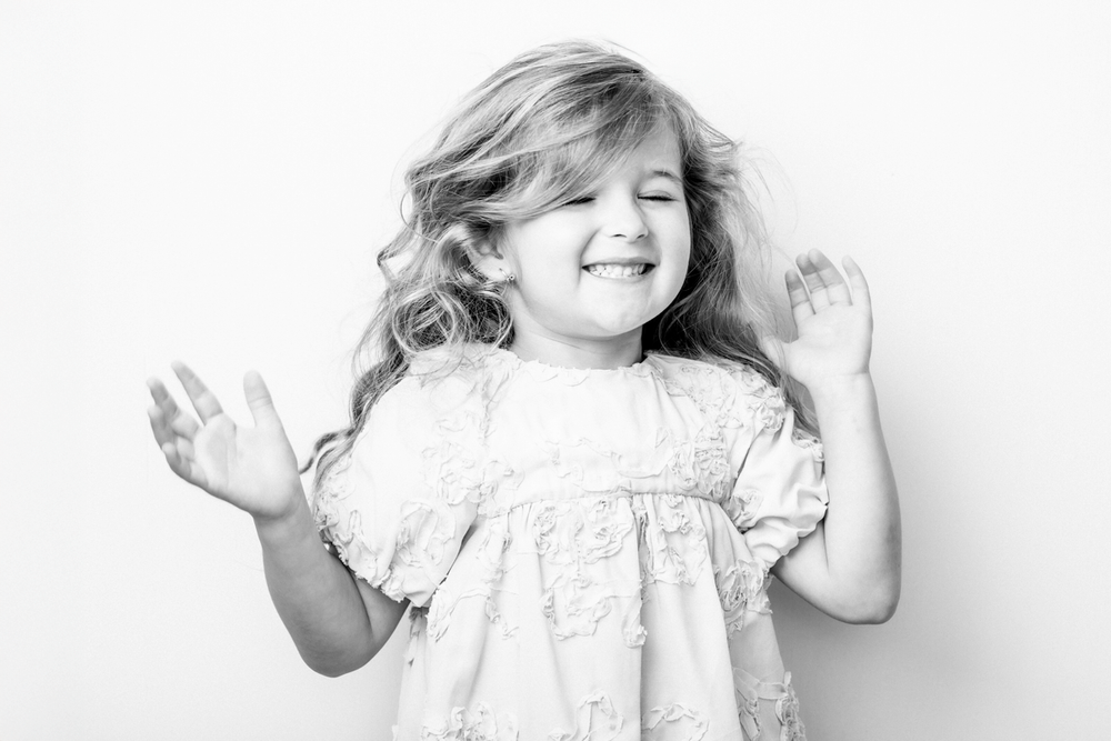 Black and white portrait of a joyful child with wavy hair wearing a white dress and raising their hands while smiling.