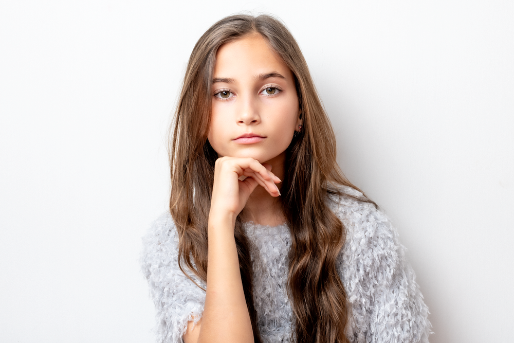Portrait of a pensive young person with long brown hair wearing a gray sweater against a white background.