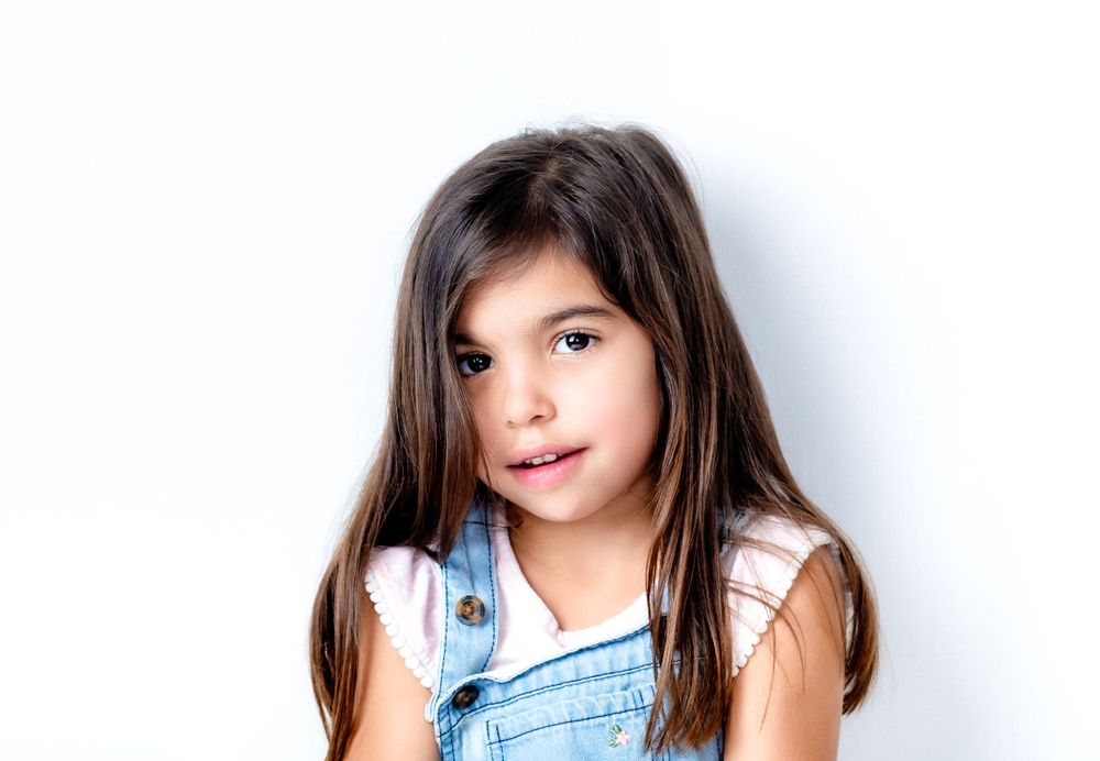 Young child with long dark hair wearing denim overalls against white background.