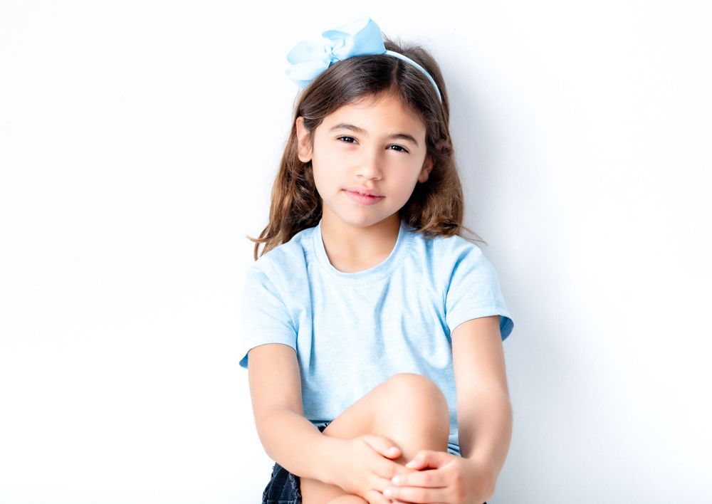 A young person in a light blue t-shirt sits against a white wall with a thoughtful expression.