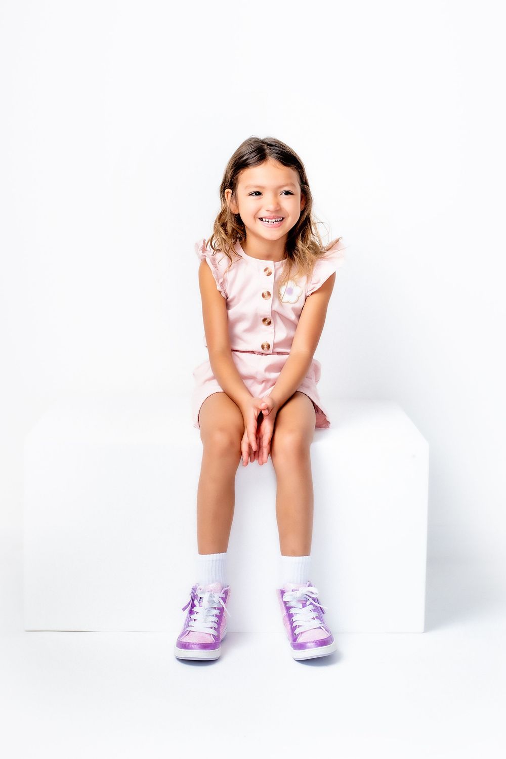 Young child in pink outfit and purple sneakers sits smiling on white block against white background.