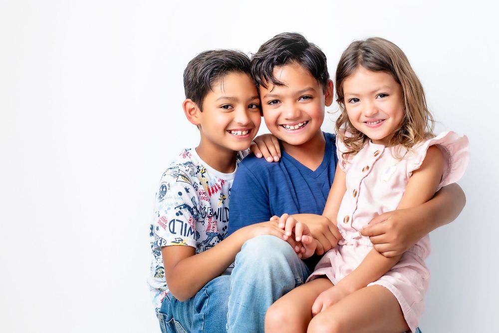 Three smiling children sitting together against a white background wearing casual clothing.