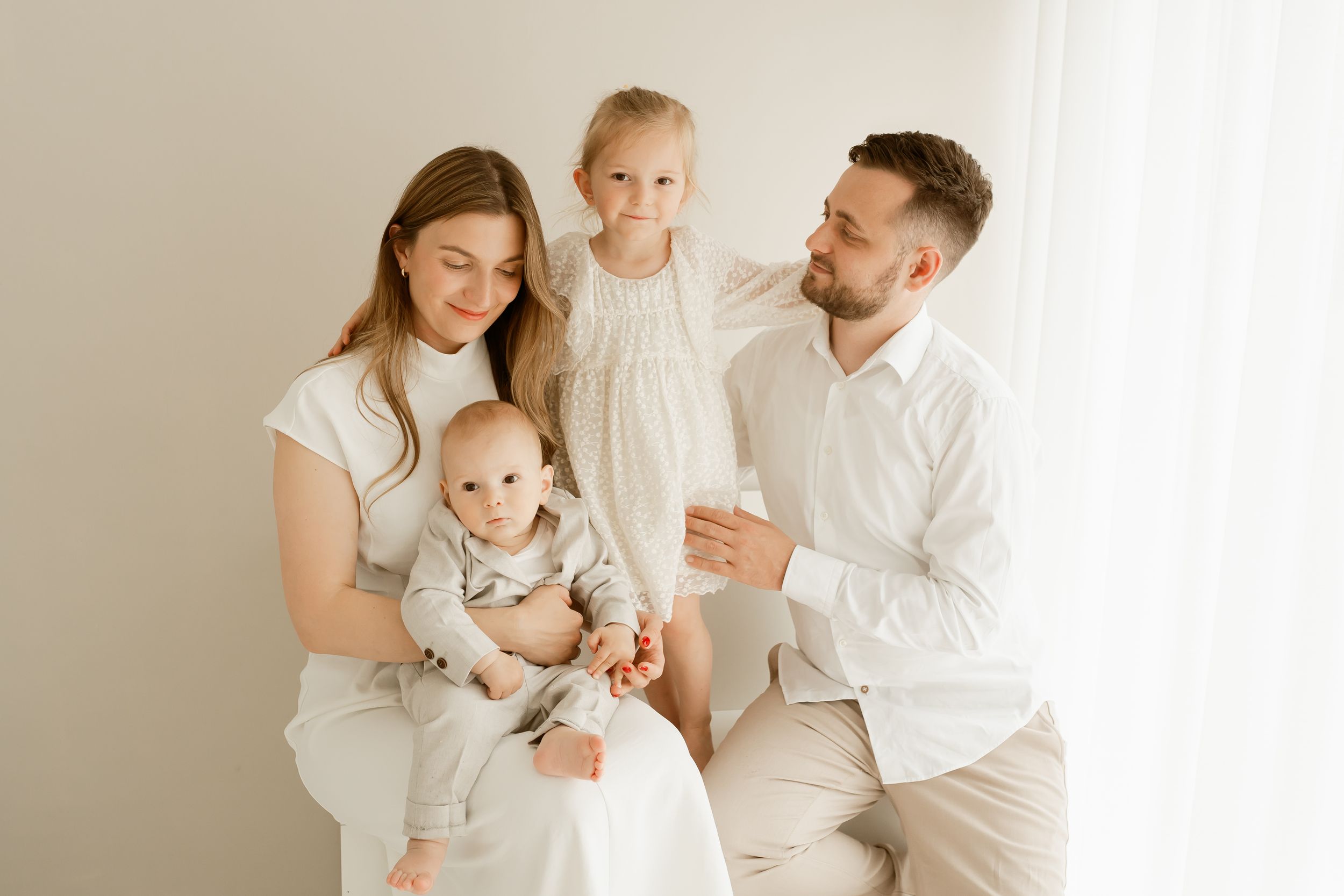 A family in white outfits shares tender moments together against a light neutral background.