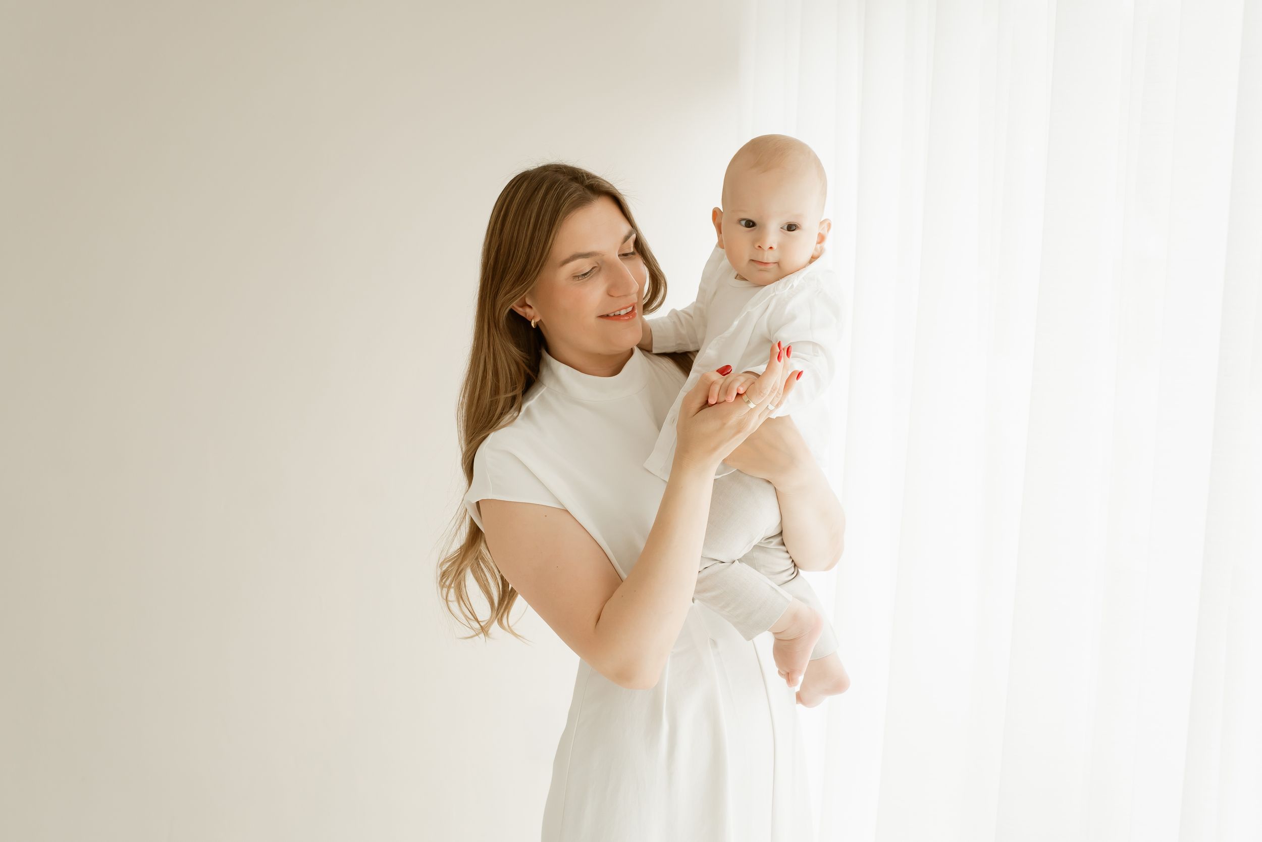 Parent and baby dressed in white outfits posing together against light neutral backgrounds in a minimalist photoshoot.