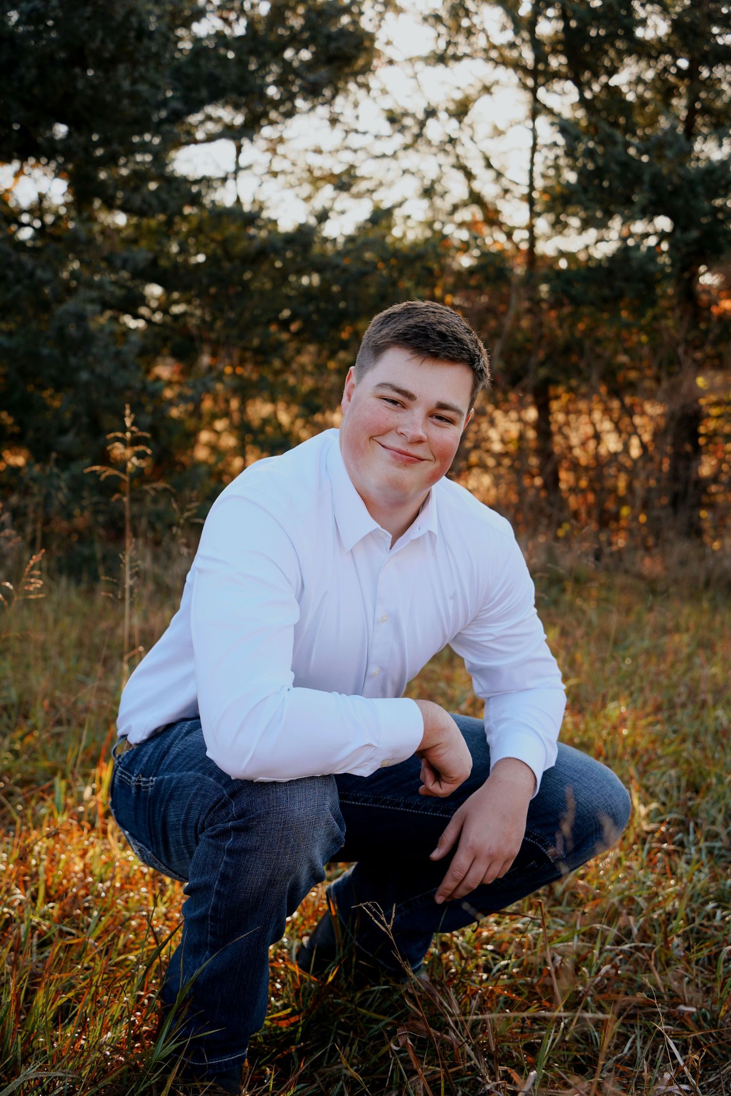 Young man in white shirt and jeans crouching in golden autumn field, smiling at camera.