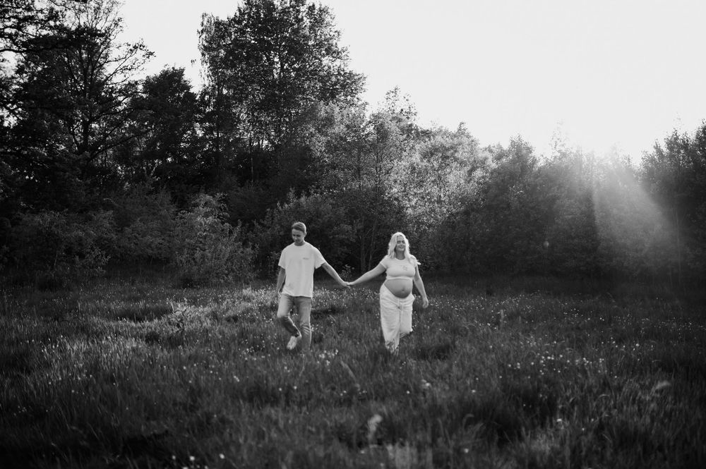 A romantic couple in white outfits holds hands while walking through a misty meadow at sunset in black and white.