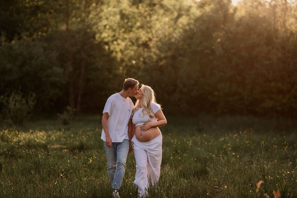 A couple in white outfits embraces while walking through a sun-drenched meadow at golden hour.