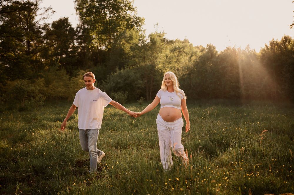 A couple holds hands while walking together through a grassy field at sunset with trees silhouetted in the distance.