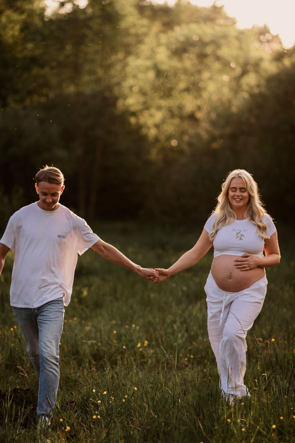 A couple holds hands while walking through a golden sun-lit meadow, with the woman showing her pregnant belly.