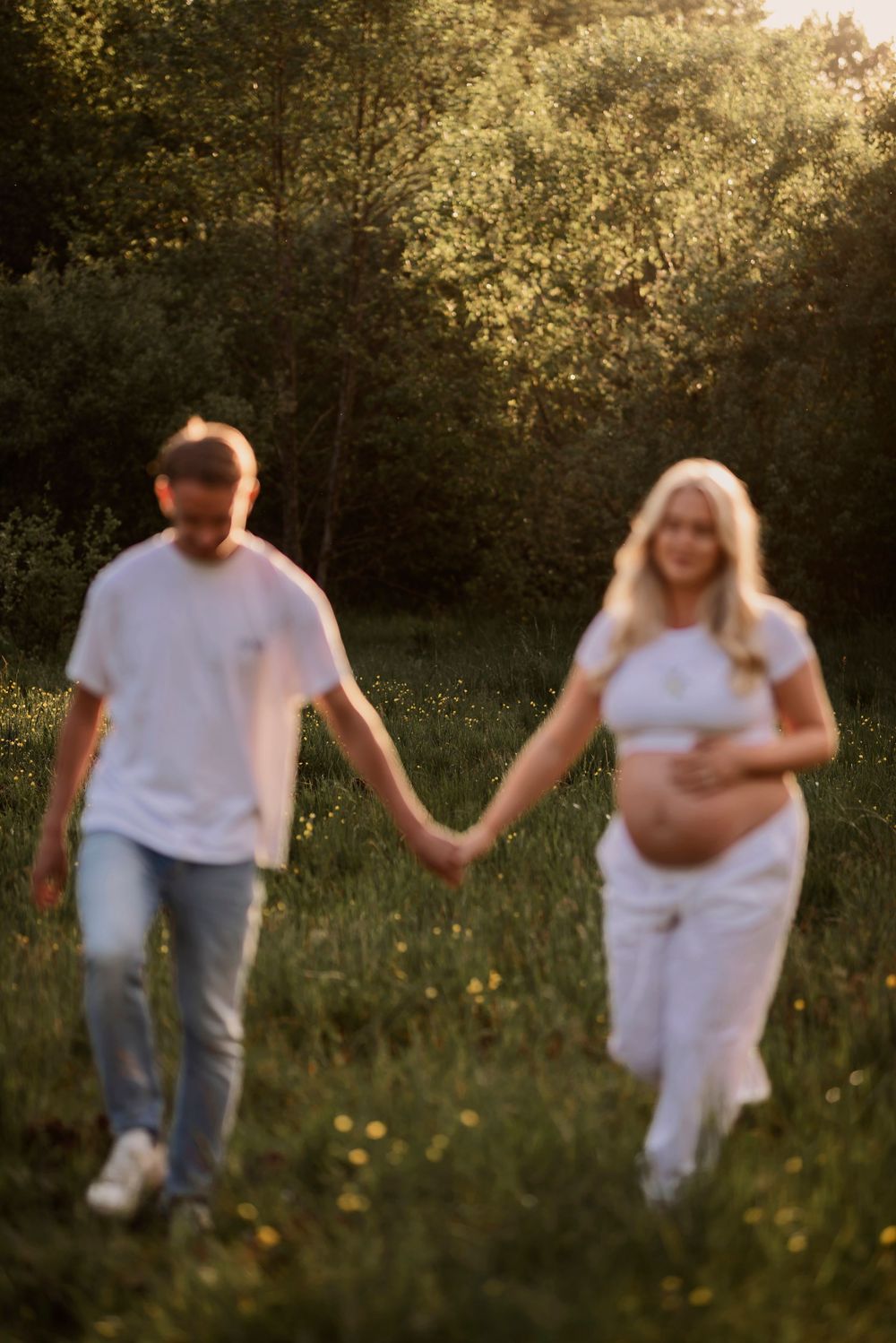 An expecting couple in white outfits walks hand-in-hand through a wildflower field at sunset.