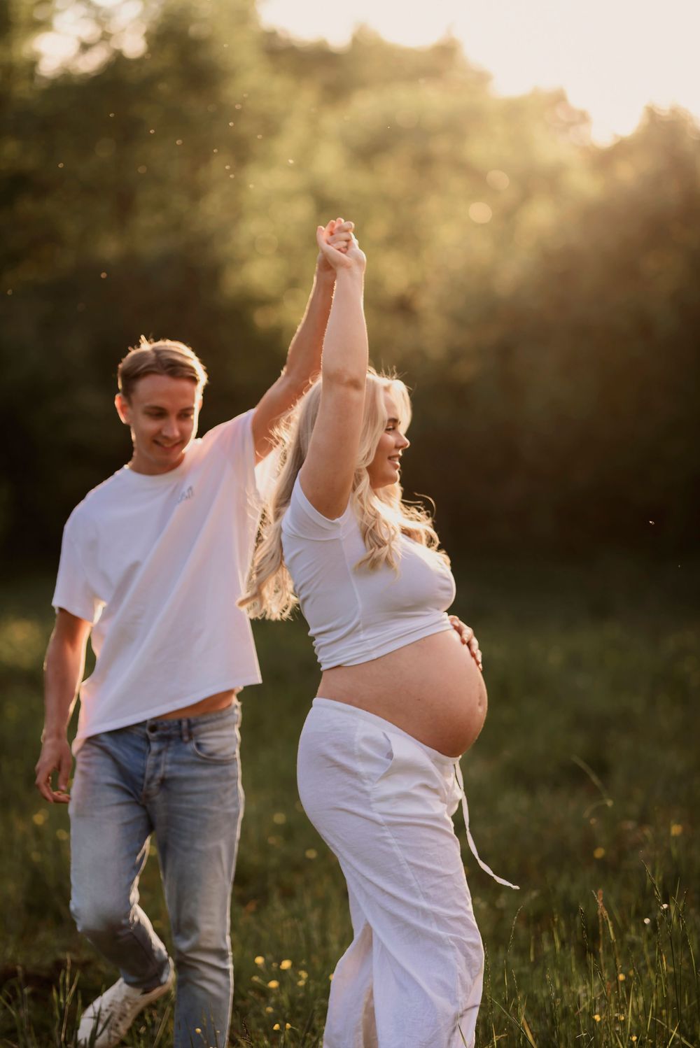 A couple in white clothing dances joyfully in a sunlit meadow during a maternity photoshoot at golden hour.