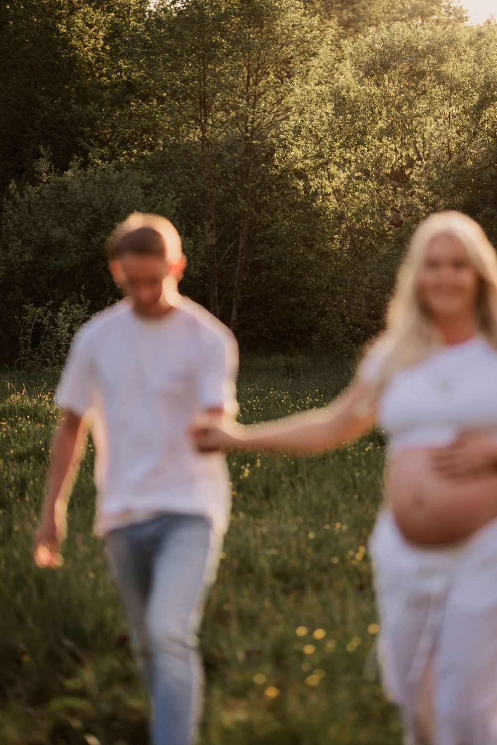 An expectant couple holds hands while walking through a grassy field at sunset.