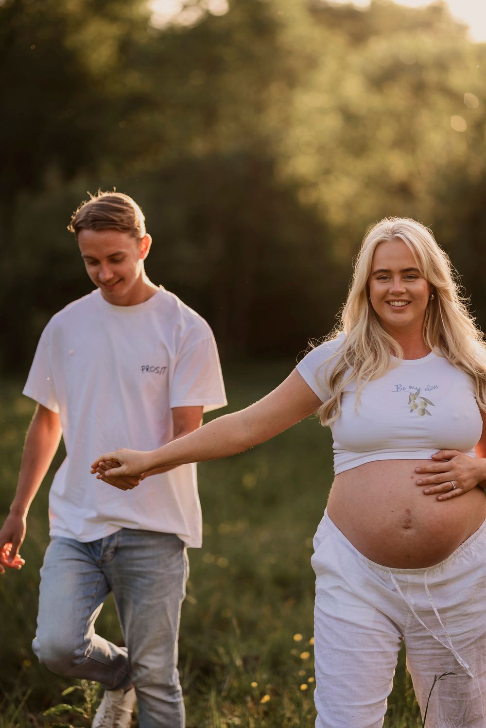 Couple poses in a sunlit meadow during pregnancy photoshoot wearing white outfits at golden hour.