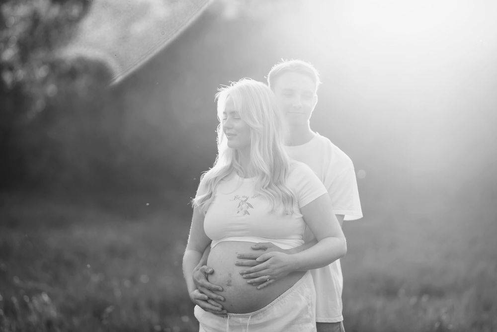 Romantic maternity photoshoot in a sunlit field shows couple embracing during golden hour.