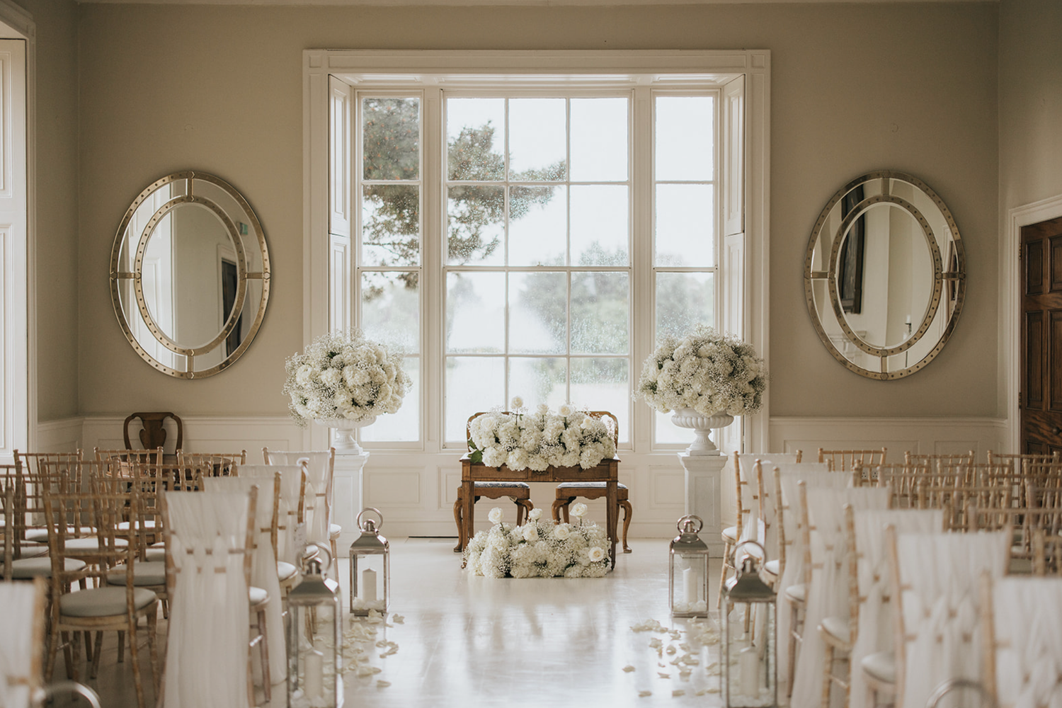 Elegant wedding ceremony setup with white chairs, floral arrangements, and two round mirrors flanking a large window.