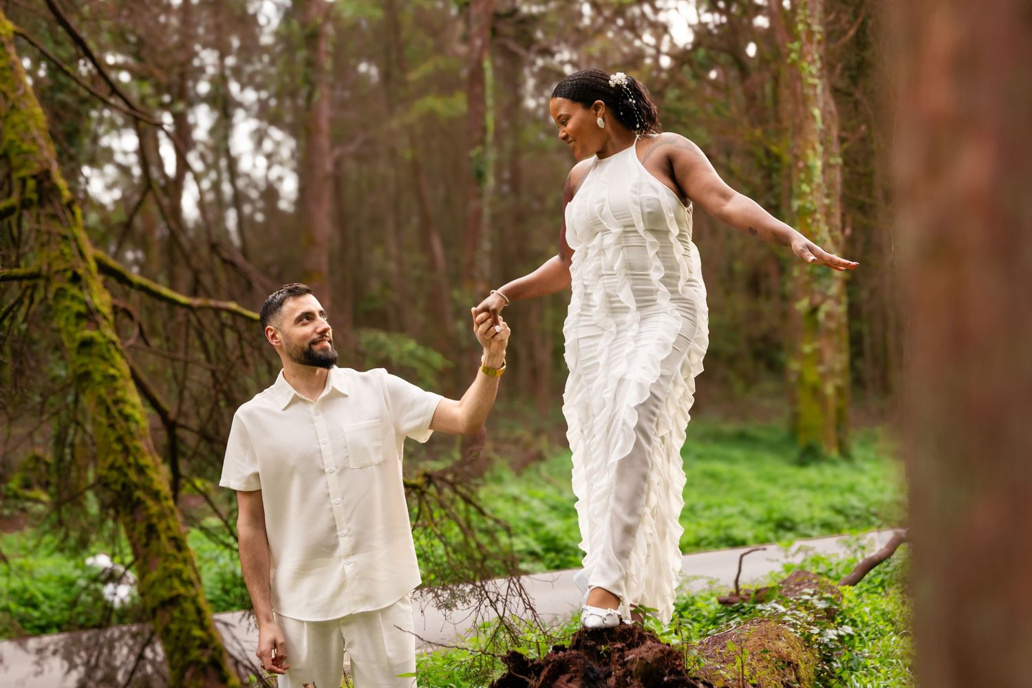 Couple in white outfits playing and dancing in a lush green forest setting.