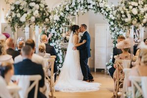 A couple stands beneath an elegant floral arch during their indoor wedding ceremony surrounded by seated guests.
