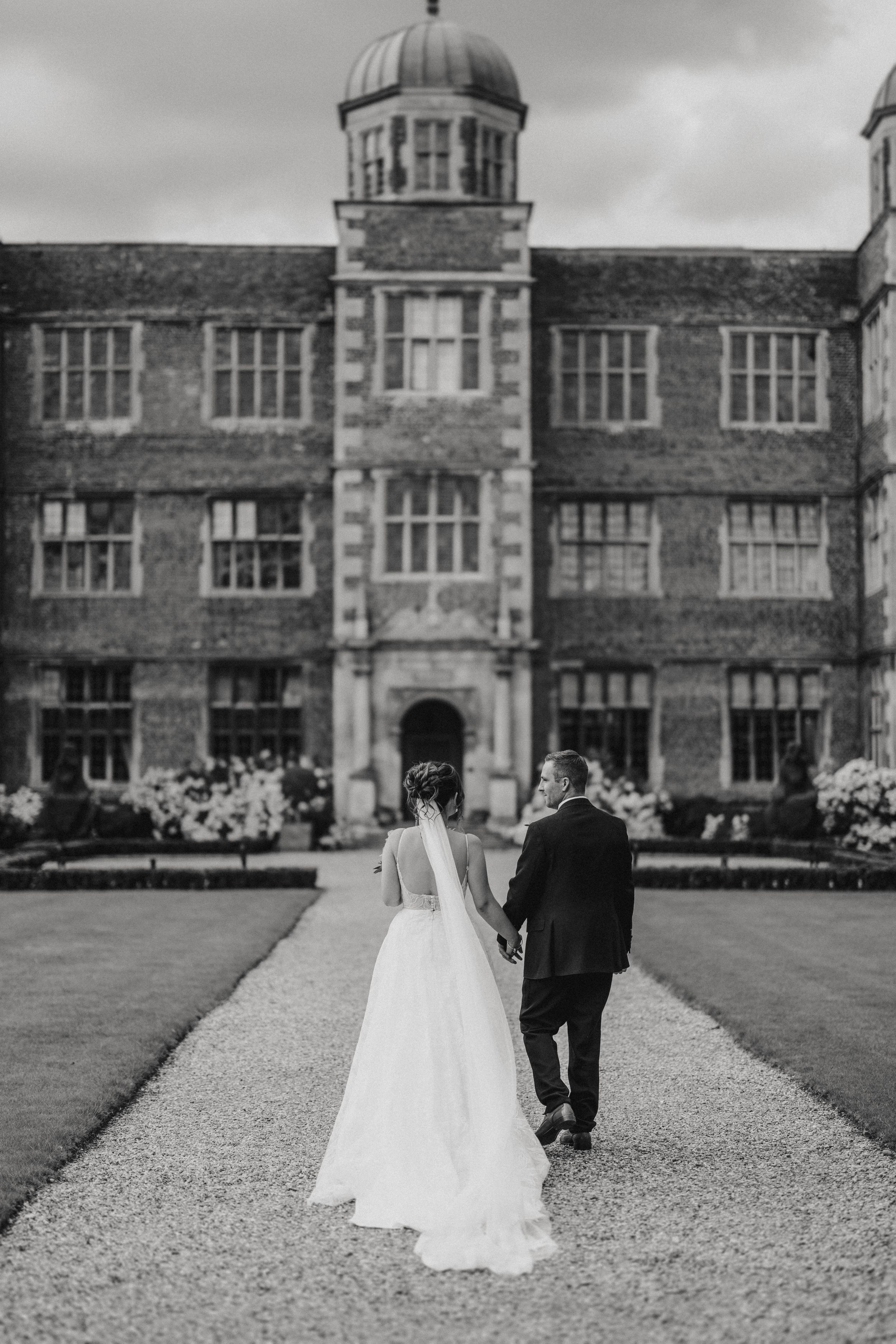 Black and white photo of a newlywed couple walking away down a gravel path toward a grand historic building with domed tower.