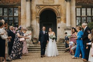 Wedding guests throw rose petals and confetti at a newlywed couple outside a historic stone church in celebration.