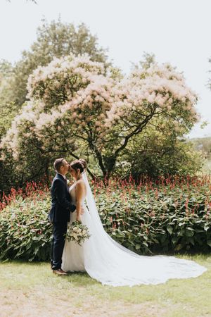 A couple in wedding attire stand together under blooming cherry blossom trees with bright red flowers in the garden.