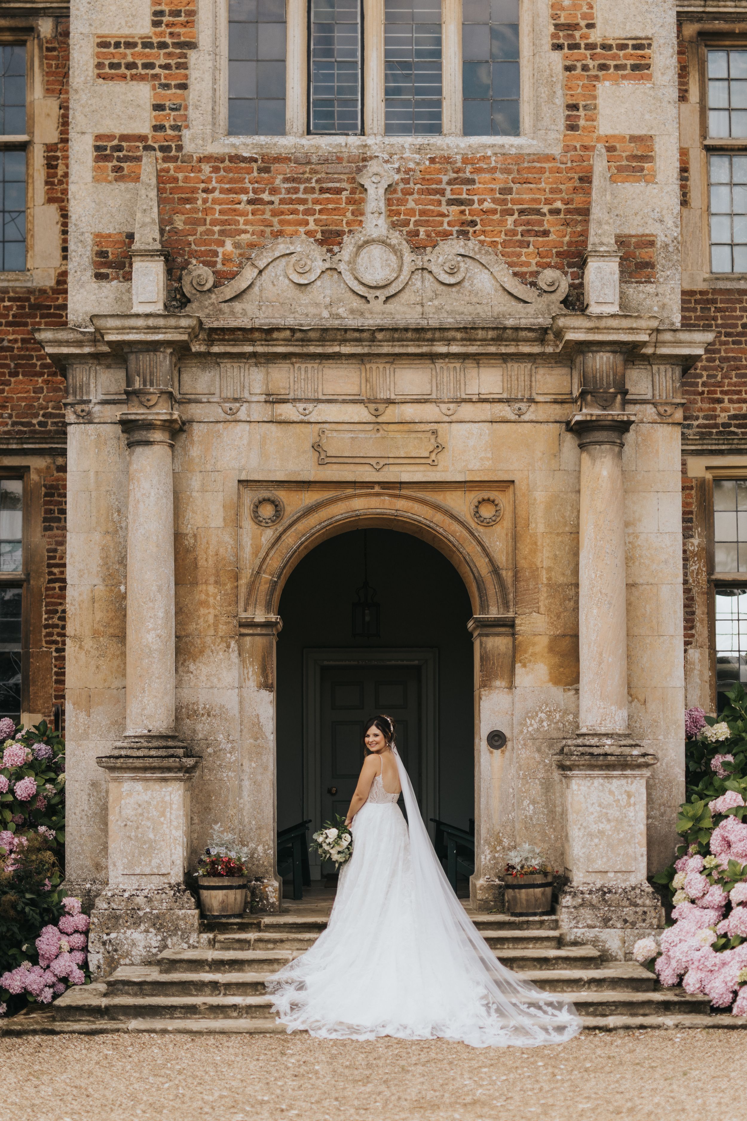 Wedding dress with long train photographed outside historic brick building with stone columns and pink flowers.