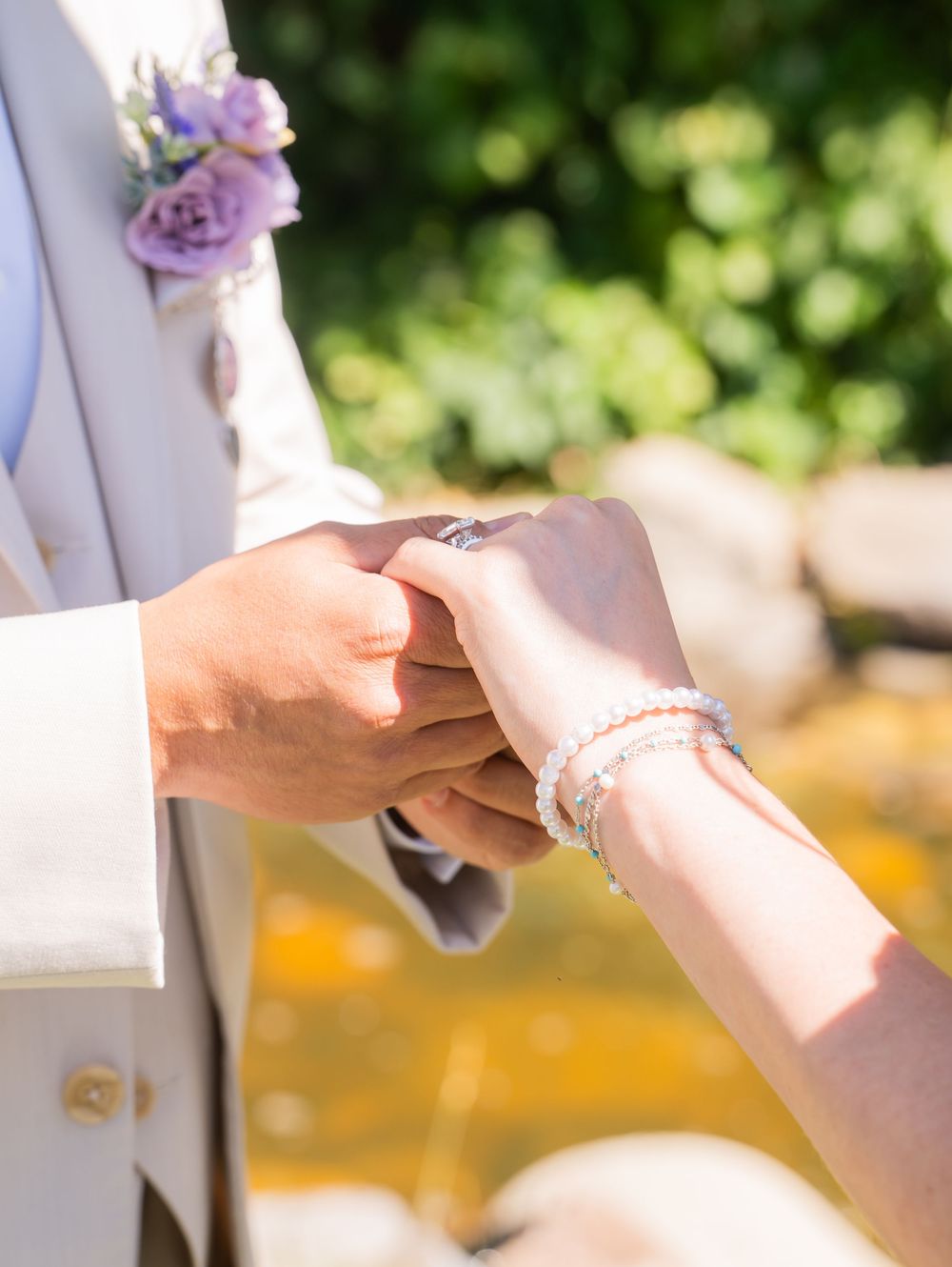Hands exchanging rings during an outdoor wedding ceremony with purple flowers and natural lighting.