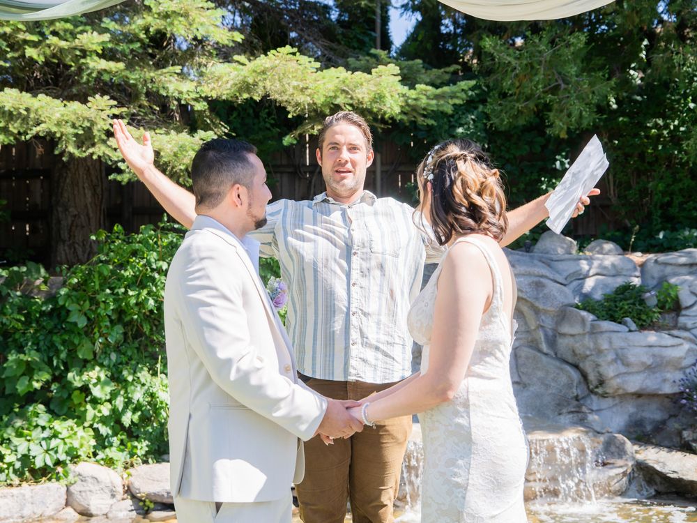 An outdoor wedding ceremony takes place in a garden setting with a cascading waterfall and lush greenery in the background.