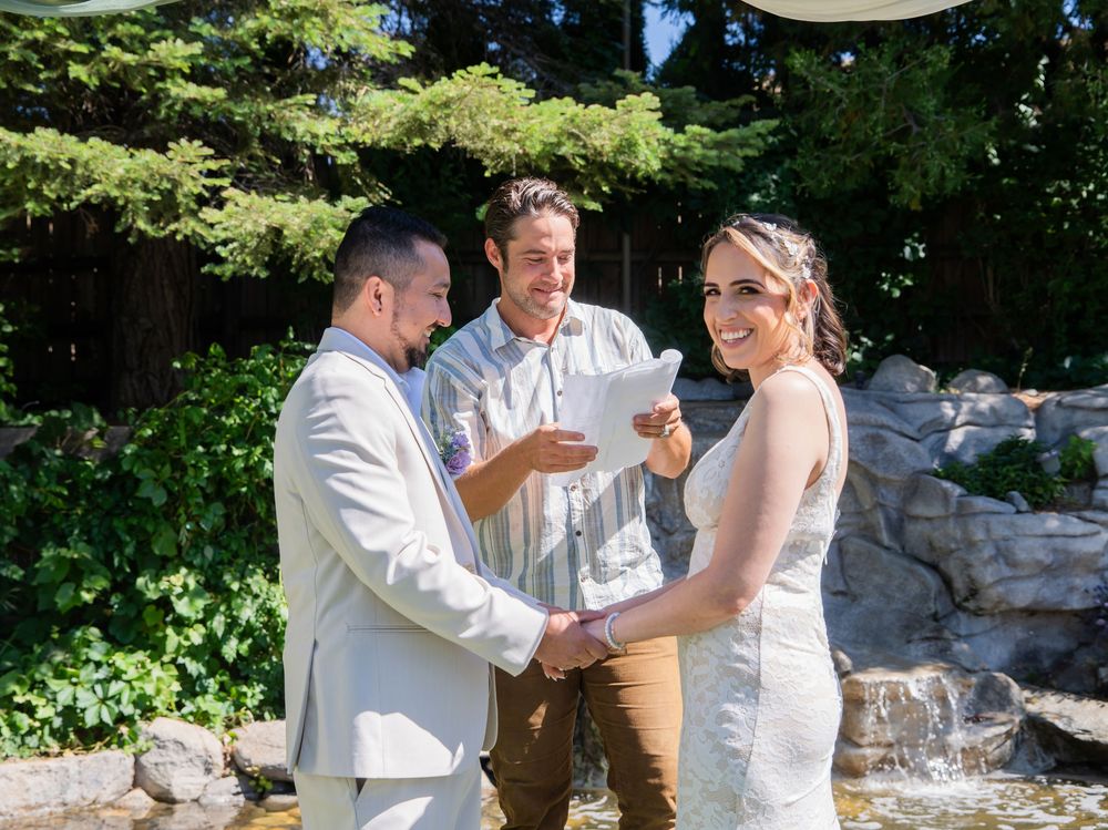 A joyful outdoor wedding ceremony takes place in a garden setting with natural rock formations and greenery in the background.