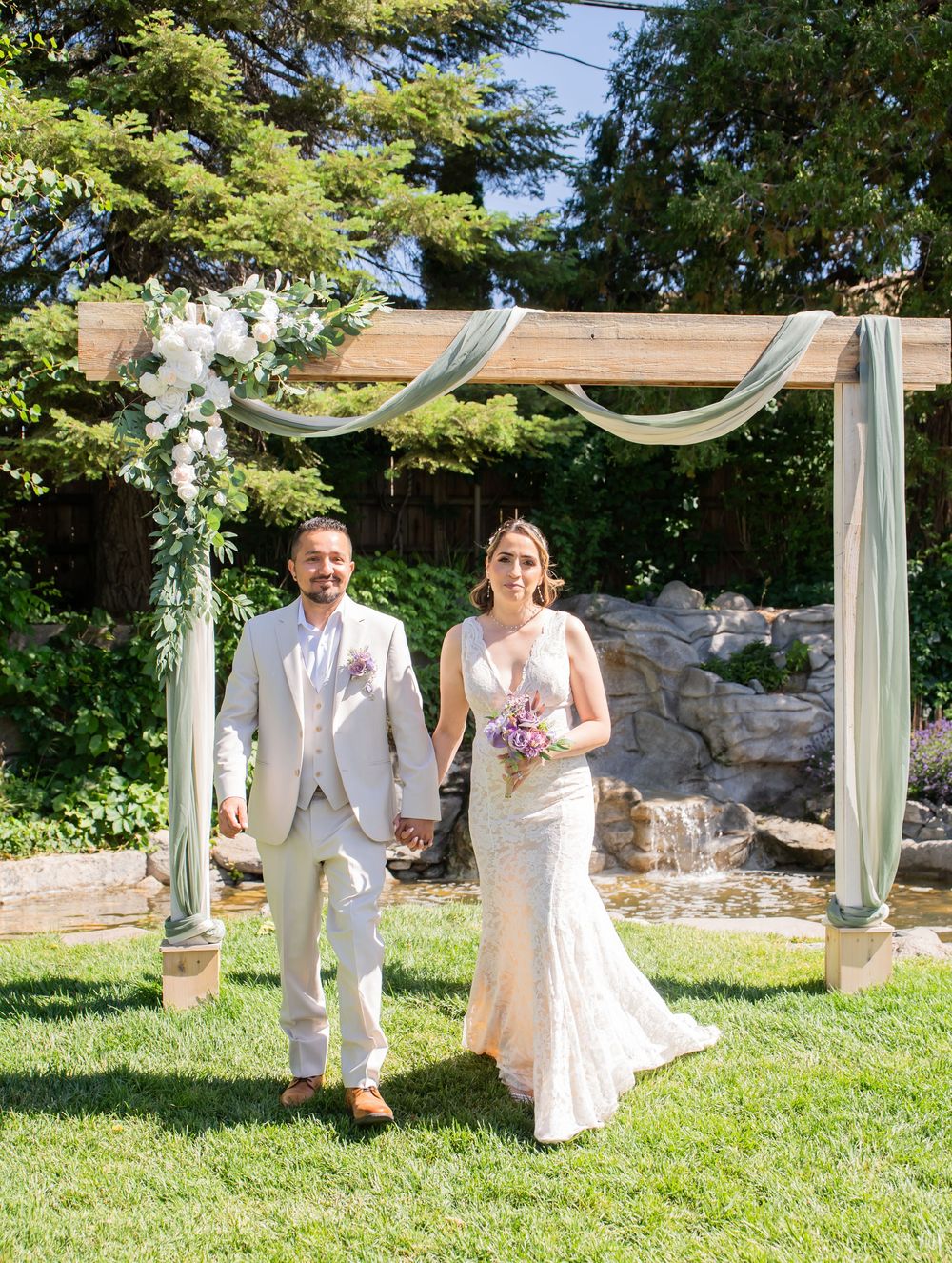 A wedding couple poses playfully under a white draped arch in a sunny garden setting.
